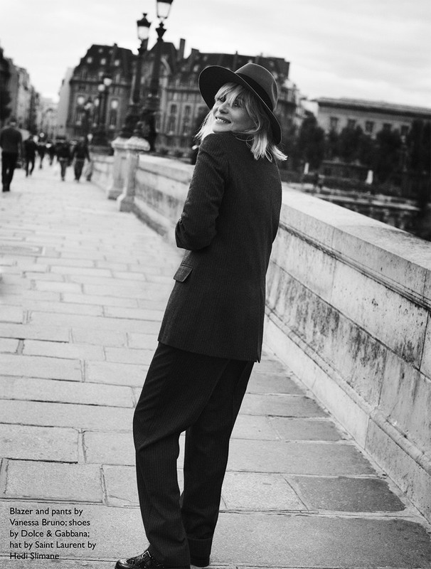 Woman in a blazer, hat, and pantsuit poses on a stone bridge with Parisian architecture visible in the background, black and white.
