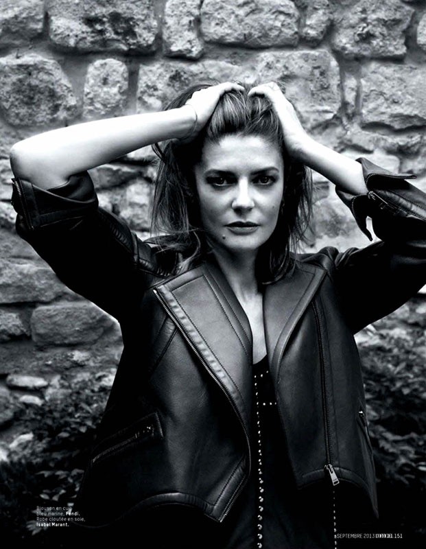 Monochrome portrait of a woman with hands in her hair, wearing a black leather jacket, against a textured stone wall.