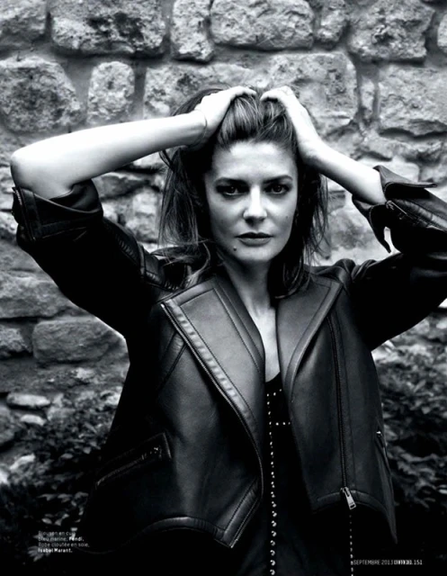 Monochrome portrait of a woman with hands in her hair, wearing a black leather jacket, against a textured stone wall.