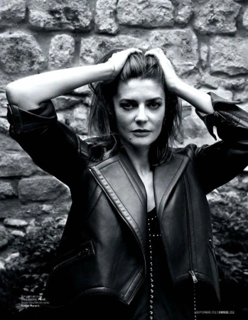Monochrome portrait of a woman with hands in her hair, wearing a black leather jacket, against a textured stone wall.