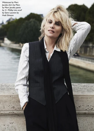 A blonde woman posing outdoors near a river wearing a pinstripe waistcoat, striped shirt, black pants, and a long black scarf.