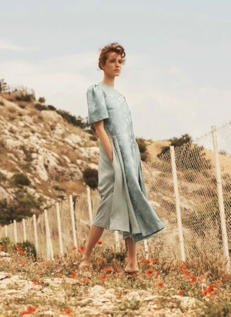 Woman in pale blue dress posing outdoors near a wire fence on a rocky hillside with scattered red wildflowers