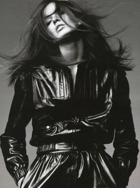 Monochrome portrait of a woman with long brown hair blowing, wearing a black leather jacket and skirt in a fashion editorial.