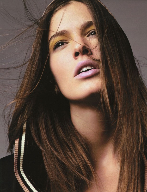 Close-up fashion portrait of a woman with long brown hair, wearing makeup, posing in front of a neutral grey background in a photo studio.