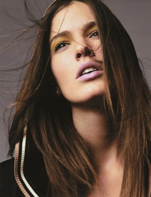 Close-up fashion portrait of a woman with long brown hair, wearing makeup, posing in front of a neutral grey background in a photo studio.