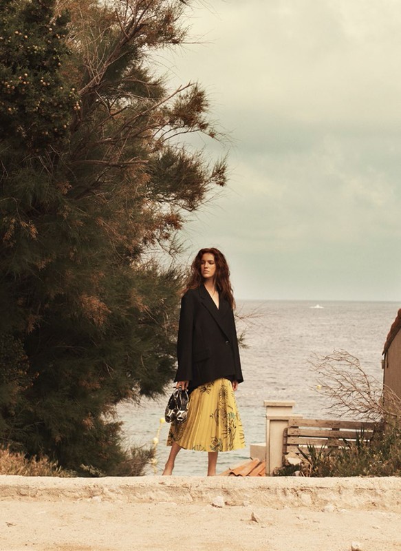 Adult woman in a black blazer and yellow skirt stands by the ocean near trees. The sky is overcast and the photo has warm tones.