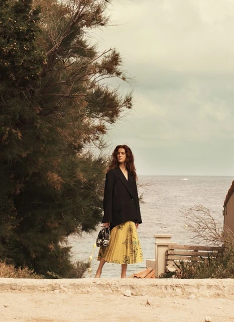 Adult woman in a black blazer and yellow skirt stands by the ocean near trees. The sky is overcast and the photo has warm tones.