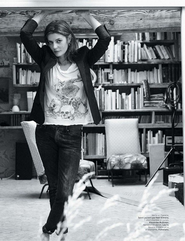 Indoor portrait of an adult woman in a t-shirt and black blazer, standing under a wooden beam with a bookshelf background.