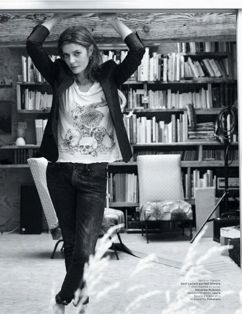 Indoor portrait of an adult woman in a t-shirt and black blazer, standing under a wooden beam with a bookshelf background.