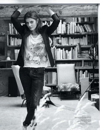 Indoor portrait of an adult woman in a t-shirt and black blazer, standing under a wooden beam with a bookshelf background.