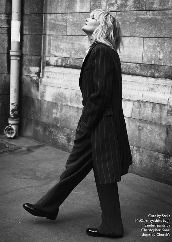 A woman in a pinstripe coat and dark pants walks along a city street in a black and white editorial fashion photograph.