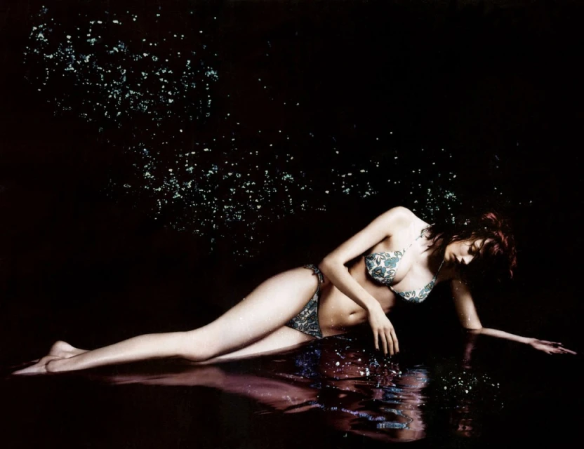 Fashion shot of a woman in a bikini lying in shallow water with glittering particles floating in the air and a dark background.