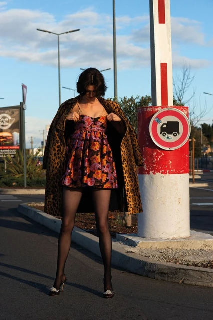 Young woman poses outdoors in a floral dress, animal print coat, and tights, standing near a red and white barrier on a sunny day.