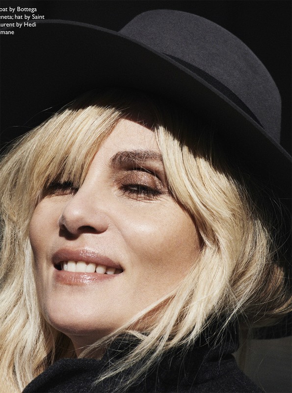 Close-up of a blonde woman in a black hat and coat, bathed in sunlight, showcasing high-fashion editorial style and beauty.