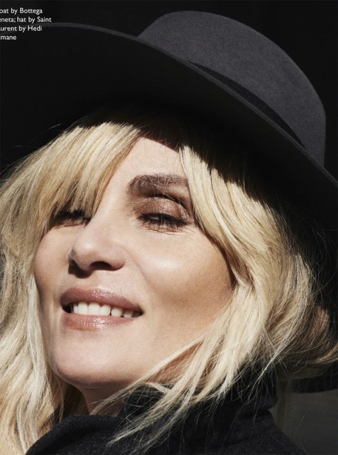 Close-up of a blonde woman in a black hat and coat, bathed in sunlight, showcasing high-fashion editorial style and beauty.