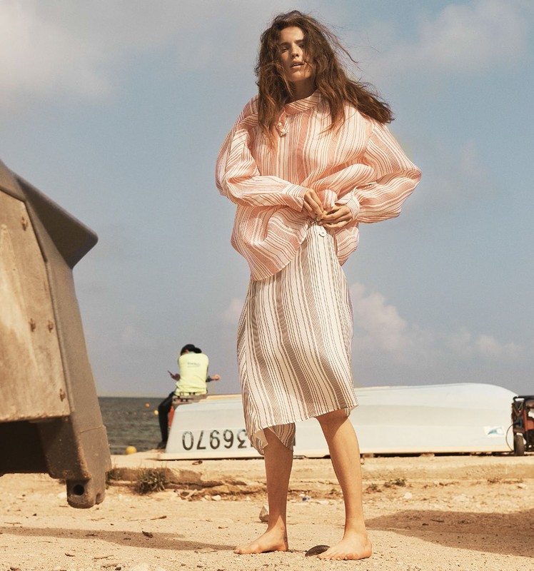 A barefoot woman in a striped blouse and skirt stands on a beach near a boat in a sun-drenched outdoor fashion shot.