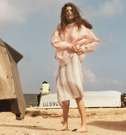 A barefoot woman in a striped blouse and skirt stands on a beach near a boat in a sun-drenched outdoor fashion shot.