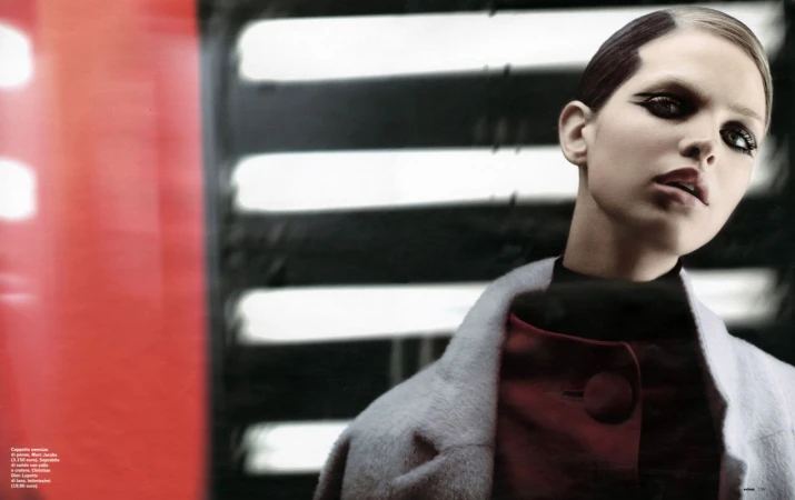 Fashion editorial shot featuring a model in red and gray clothing with dramatic eye makeup, posing against a blurred backdrop.