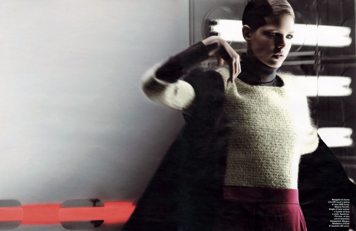 A young woman poses indoors in a neutral sweater and maroon skirt. The photo features motion blur, red light bars, and soft studio lighting.
