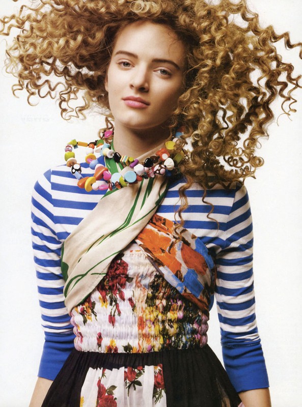 Stylish young woman with curly hair and colorful necklace wearing a striped top and floral dress against a neutral background.