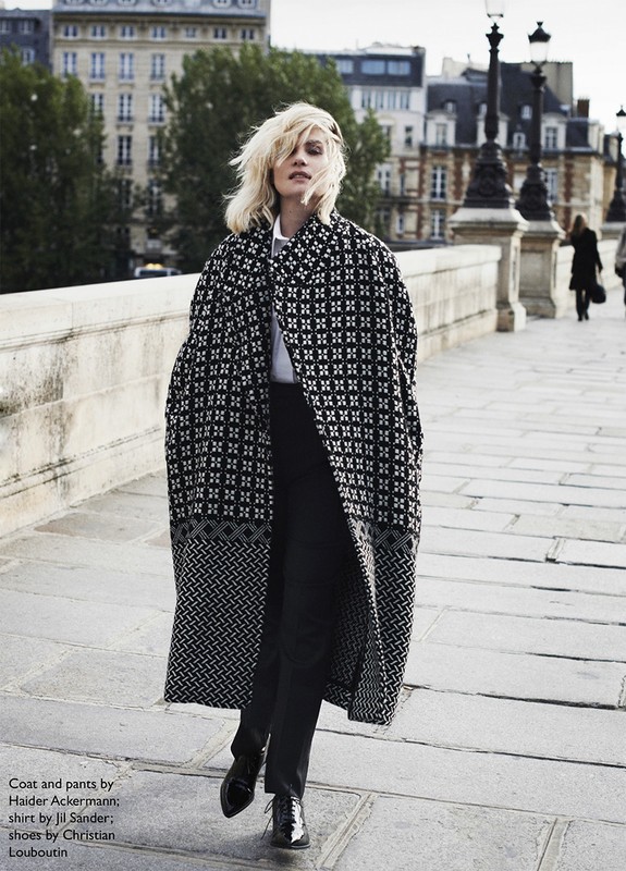 Fashion editorial shot of a blonde woman walking on a bridge in Paris, wearing a long patterned coat, black pants, and glossy dress shoes.