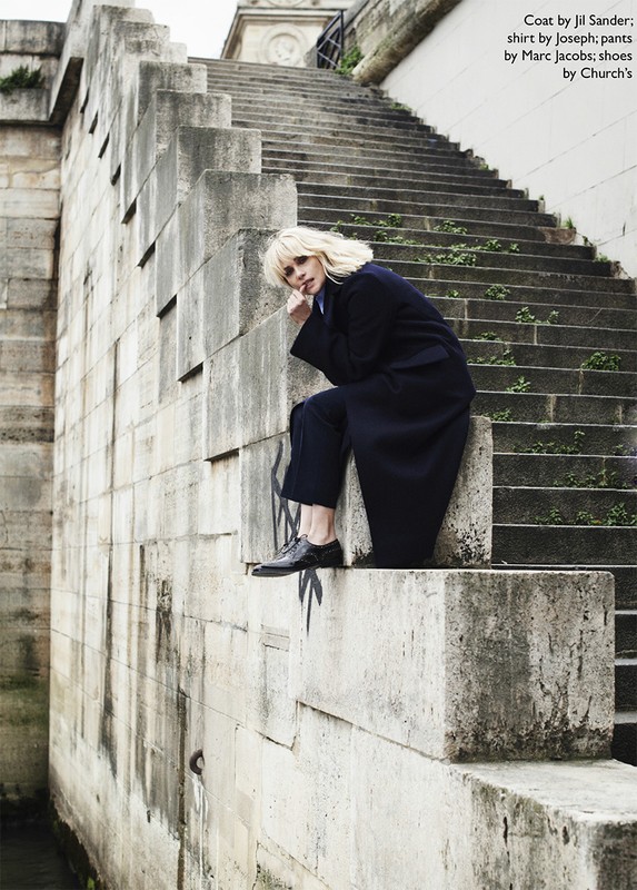 Fashion editorial image featuring a woman in a dark coat and pants sitting on stone steps outdoors in an urban environment.