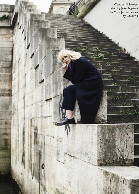 Fashion editorial image featuring a woman in a dark coat and pants sitting on stone steps outdoors in an urban environment.
