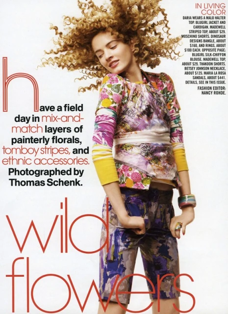 Fashion editorial shot of a woman with curly hair wearing a floral top, shorts, and accessories, against a white background. Magazine advertisement.