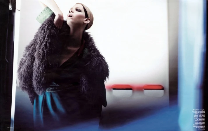 Model wearing a furry dark vest and green top posing dynamically against a minimal white wall with red details.