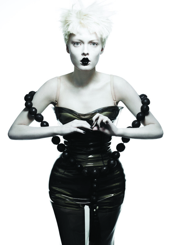 A woman with short blonde hair, pale skin, and black lipstick poses in a glossy black dress and beaded necklace against a white background.