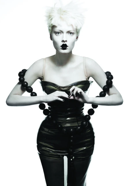 A woman with short blonde hair, pale skin, and black lipstick poses in a glossy black dress and beaded necklace against a white background.