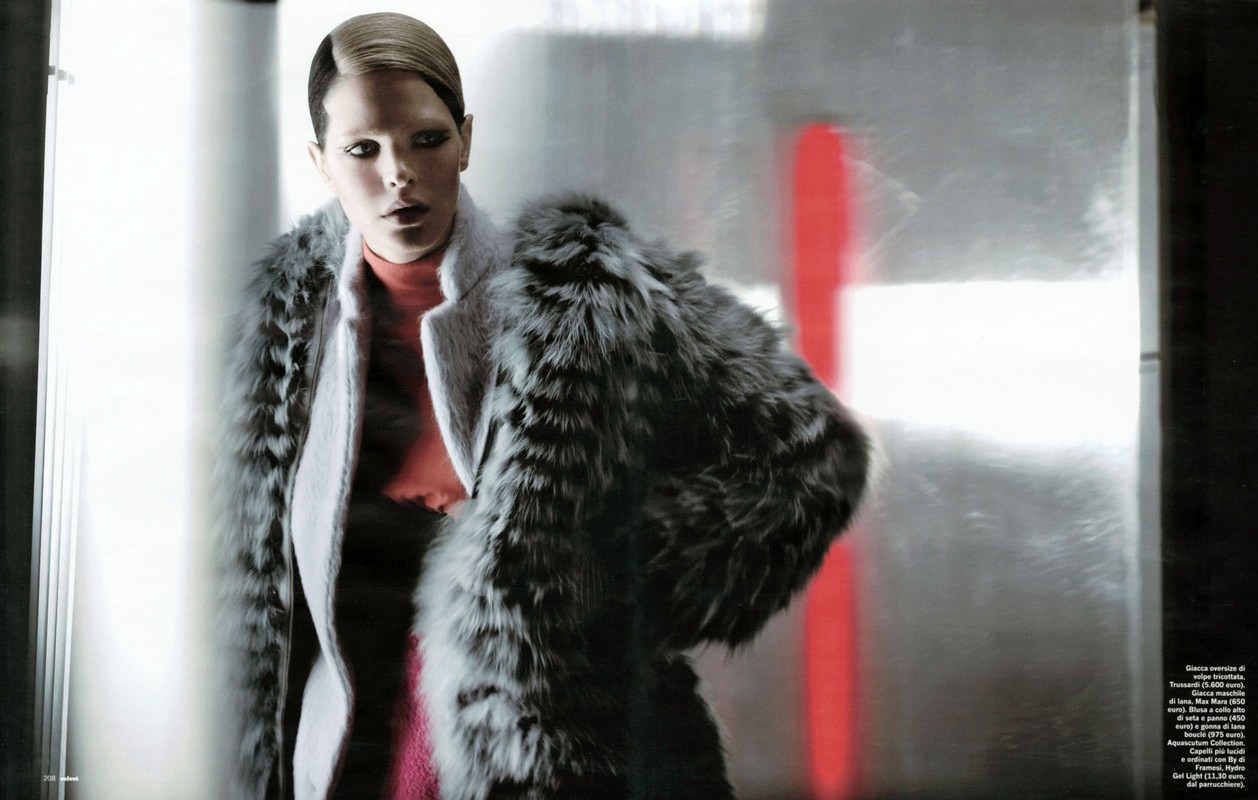 Fashion editorial shot of a female model with fair skin, wearing a grey coat and a fur stole, posed in an indoor setting with diffused light.