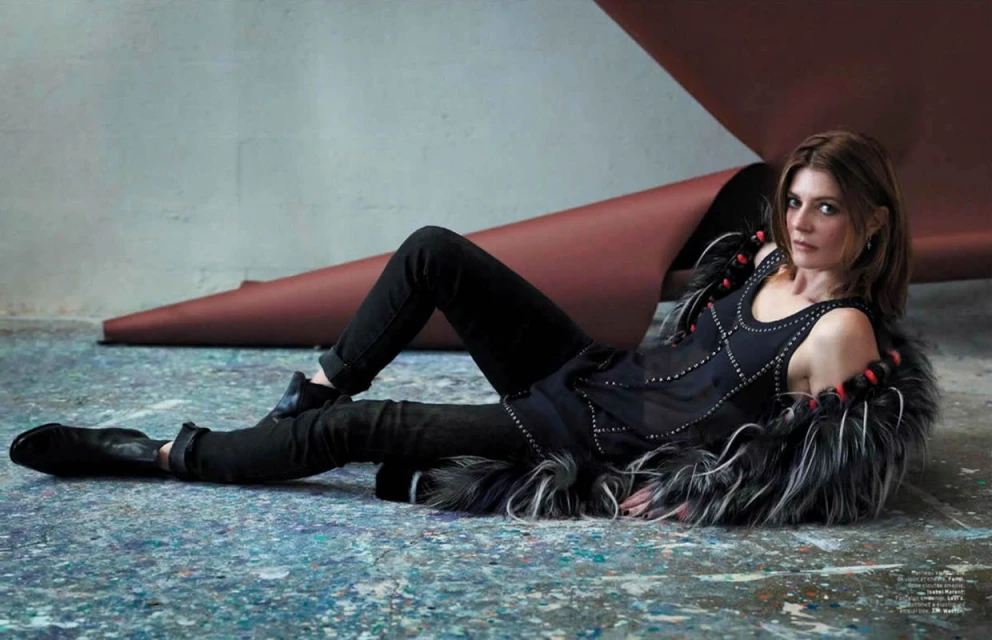 A woman in a black top and jeans poses while laying on a paint-splattered surface, wearing a grey fur stole, indoors.