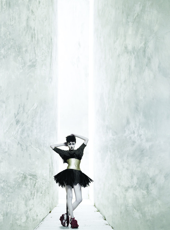 Fashionable woman strikes a pose in an all-black dress with a gold corset and red heels, standing in a long corridor.