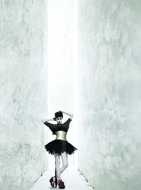 Fashionable woman strikes a pose in an all-black dress with a gold corset and red heels, standing in a long corridor.