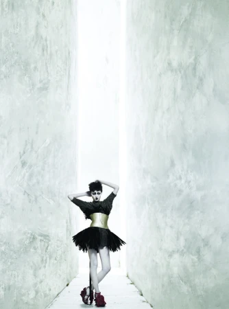 Fashionable woman strikes a pose in an all-black dress with a gold corset and red heels, standing in a long corridor.