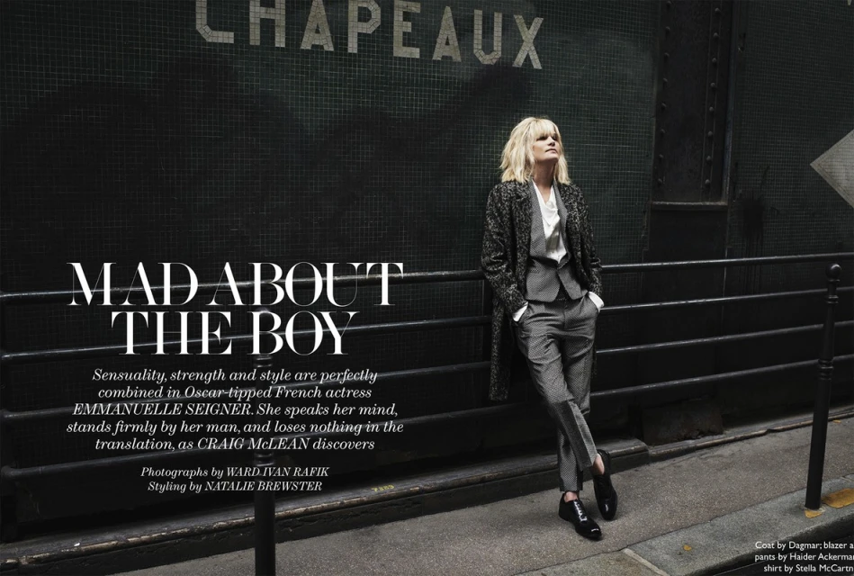 Stylish woman in grey suit and patterned coat leans against a dark wall with Parisian text. Outdoor fashion editorial shoot.