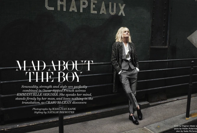 Stylish woman in grey suit and patterned coat leans against a dark wall with Parisian text. Outdoor fashion editorial shoot.
