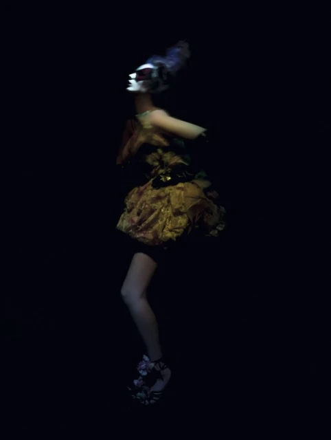 Fashion shot of an adult female model wearing a couture dress with floral detail and a masquerade mask in a dark studio setting.