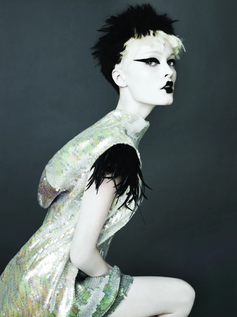 Fashionable person with dramatic black and white hair and makeup wears a shimmering iridescent sequined dress with black feathers.