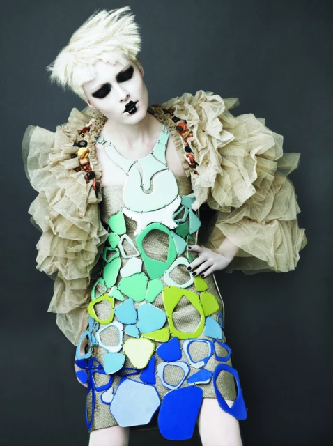 Blonde woman in a sculptural fashion dress with a beige ruffled shawl poses in a studio setting with dramatic dark eye makeup.