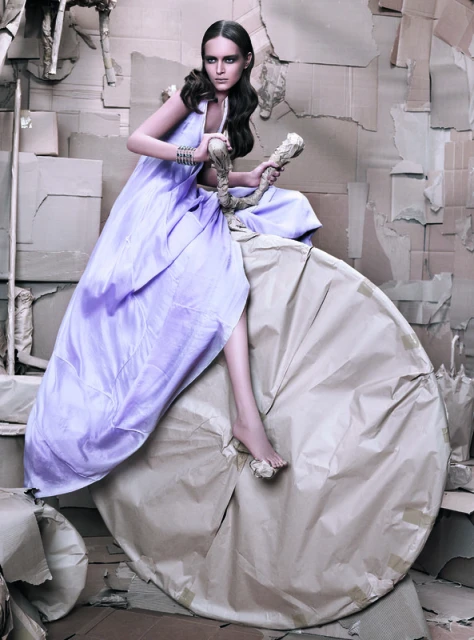 Glamour shot of an adult female fashion model in a purple dress, seated on a large cardboard roll, set against a cardboard background.