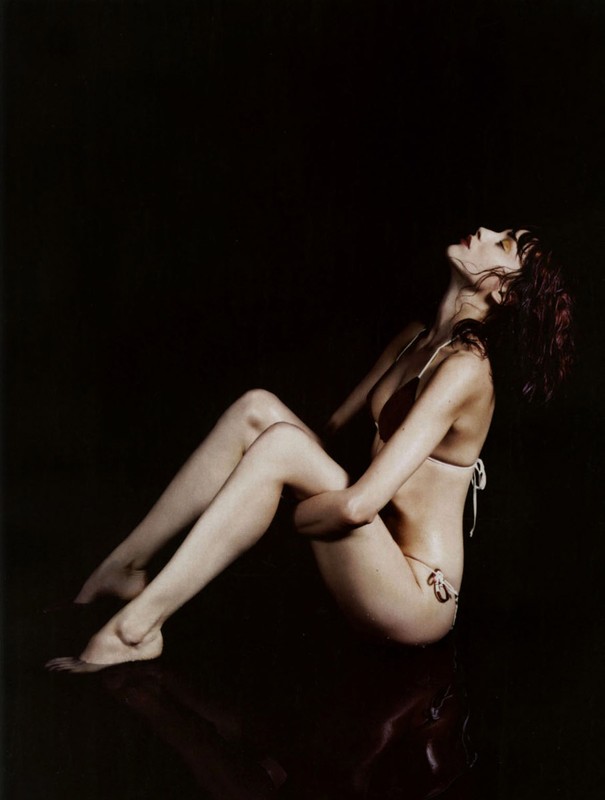 Woman in bikini sitting with head back in dark studio. Female fashion portrait with skin and body tone contrast against black background.