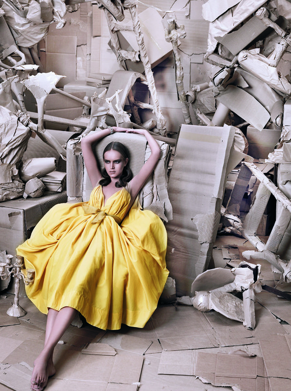 Fashion shot of a young adult female model in a yellow dress, seated in a chair surrounded by a cardboard installation in an indoor setting.