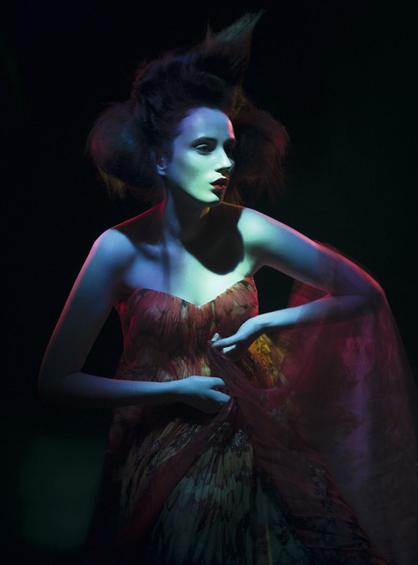 A woman with stylized hair poses in a strapless dress under blue and pink lighting against a dark backdrop in a fashion-style shot.