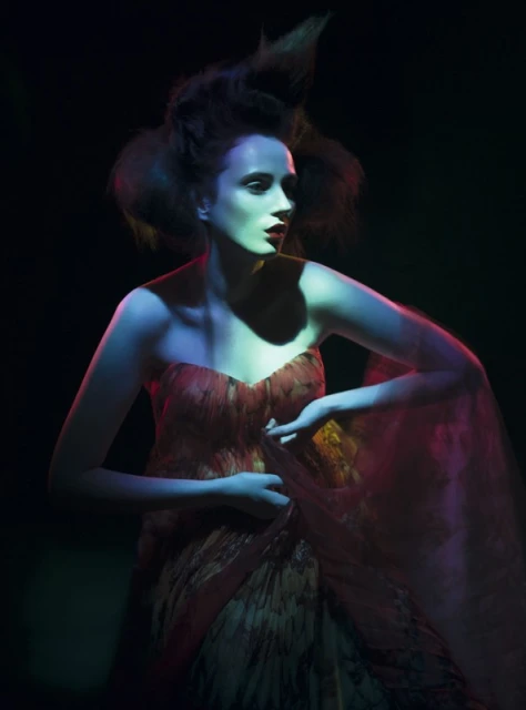 A woman with stylized hair poses in a strapless dress under blue and pink lighting against a dark backdrop in a fashion-style shot.