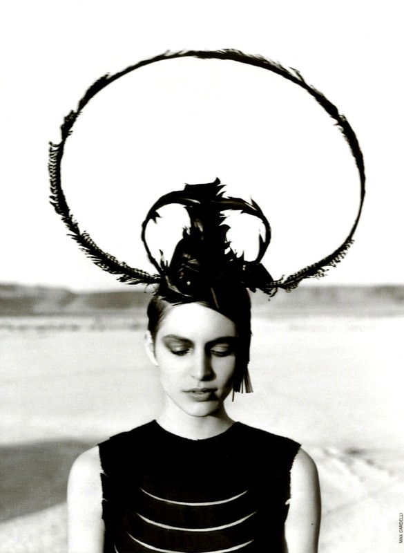 Monochrome portrait of a young woman with a feathered headdress against a soft, blurred desert backdrop, in a high-fashion editorial style.