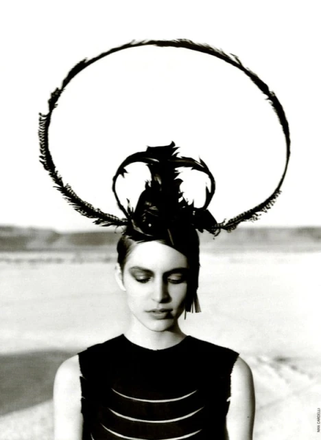 Monochrome portrait of a young woman with a feathered headdress against a soft, blurred desert backdrop, in a high-fashion editorial style.