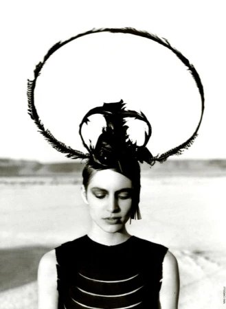 Monochrome portrait of a young woman with a feathered headdress against a soft, blurred desert backdrop, in a high-fashion editorial style.