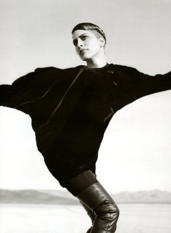 Black and white fashion shot of a slender adult woman in an oversized black blouse and leather boots posing in an open outdoor area.